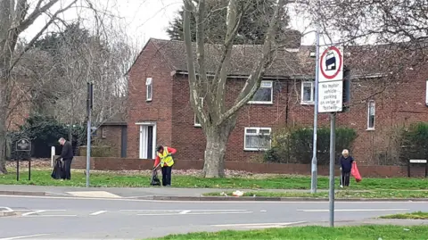 Mark Fishpool Three people collecting rubbish and litter in a residential street in Peterborough. There is housing behind them and grass verges and trees near the road junction. One person is wearing a high-visibility yellow tabard and the others are in dark clothing. 