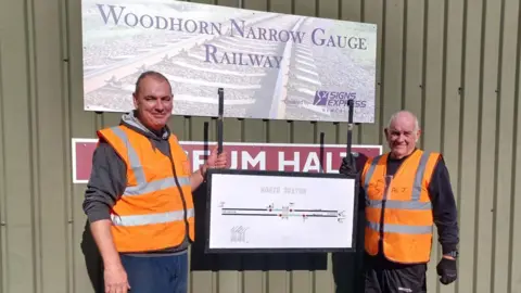 Two members of the Woodhorn Narrow Gauge Railway holding a diagram of the North Seaton signal box. The diagram shows to lines representing tracks and the signal box. The men are wearing orange hi-vis jackets.