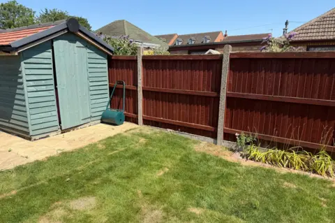 The corner of a garden with a green lawn, which looks as if it has been relaid, a brown fence and a green wooden shed.