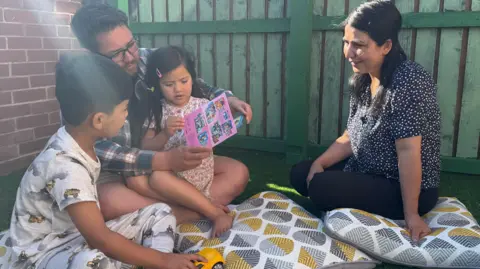 Hossain and Fereshta are sat in their garden, playing and reading with two of their children. 