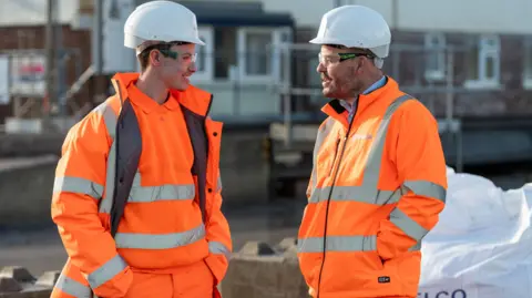 Sibelco A young man wearing bright orange safety clothing talks to an older man dressed the same way, both wearing hard hats 