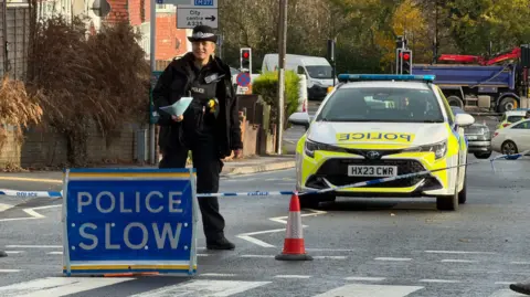 A police cordon with a Police Slow sign, blue tape, a police officer and a police car blocking the road. A pedestrian crossing can be seen in the foreground and with other vehicles including lorries in the background.