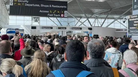 Paul Duggan A packed crowd of people in the terminal building at London Stansted Airport. There are signs above directing to "departures".