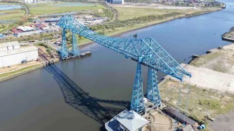 An aerial view of the blue Tees Transporter bridge over the River Tees. Fields and industrial buildings are on both riverbanks.