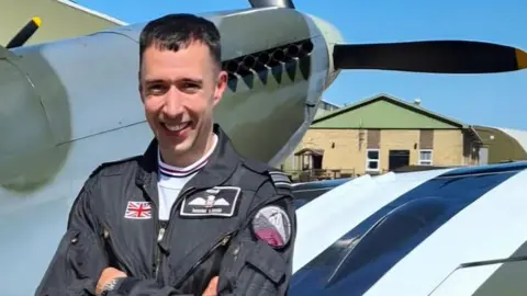 Sqn Ldr Mark Long in dark blue RAF flying suit standing with arms folded in front of a Spitfire aircraft painted grey and green with black propellers and black-and-white stripes on the wing. 