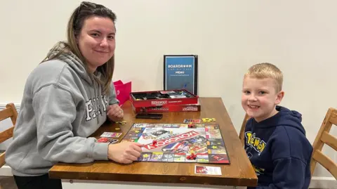 Claire is wearing a grey hoodie and is sitting at a wooden table playing a game of Monopoly with her son Riley. Riley is wearing a blue Pokemon hoodie and has blonde short hair. 