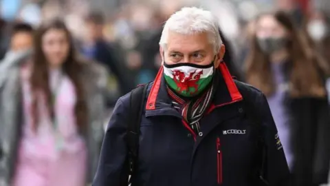 Man stood in crowded street wearing a mask with a Welsh flag on it. He is wearing a blue coat and has a rucksack on. People can be seen out of focus behind him.