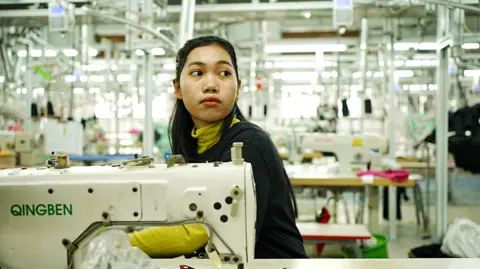 Xiqing Wang/BBC A female worker in a brightly lit factory sits behind a sewing machine. Behind her, more sewing machines can be seen. 