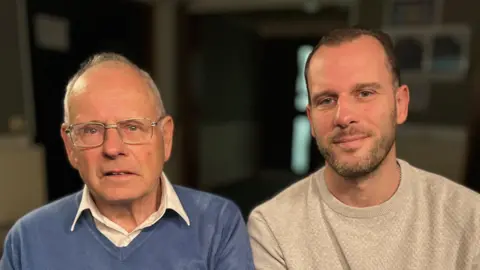 Graham Hall and Steven Hall sit next to each other and smile at the camera. Graham has thin grey hair and wears a blue jumper with a white shirt underneath. Steven has short dark hair and is wearing a cream coloured jumper.