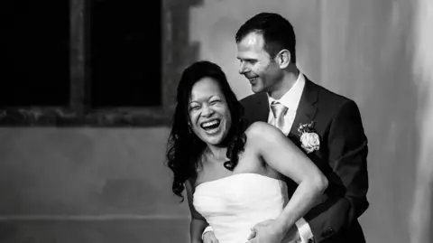 A man in a suit stood behind a woman in a white wedding dress. They are both laughing