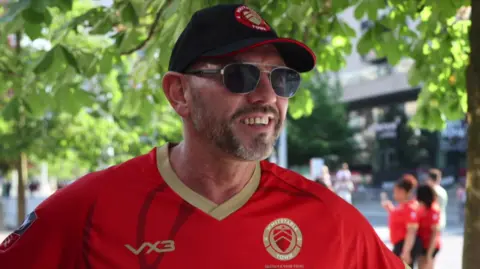 Jack Valpy/BBC, a man with a beard and sunglasses who wears a black white city FC hat, sunglasses and a red football shirt.