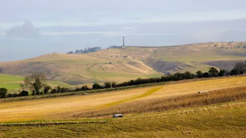 Getty Images The obelisk seen at a distance, atop brown and green hills. Brown fields are in the foreground.