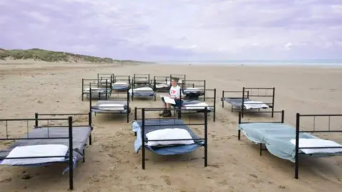 Liz Seymour Black metal bed frames lined up on the beach. The frames have been scattered around the beach.