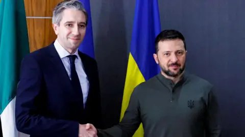 Reuters Simon Harris in a dark suit shakes hands with Volodymyr Zelensky who is wearing a green jumper. The Irish tricolour and Ukrainian flag are in the background.