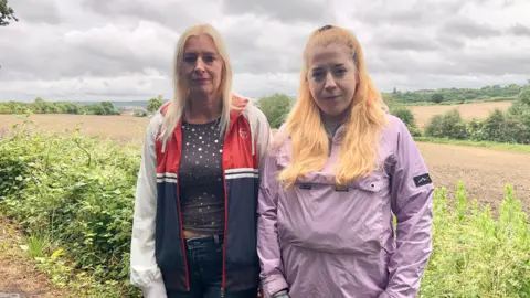 Two women are stood in front of green fields. The one on the left has shoulder length blond hair and is wearing a red, white and blue jacket with a grey spotted top and jeans. The one on the right has long strawberry blond hair and is wearing a lilac jacket