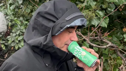 Man in black rain coat with hood up walks past shrubbery. A green drinks can is held next to his mouth. This photo is taken close up.
