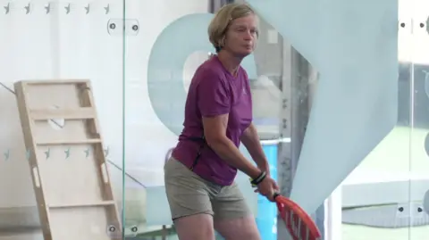 A woman holds a red padel bat as she waits to be served a ball.