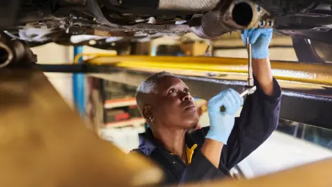 Getty Images Young black woman stands underneath car, using a tool to fix the undercarriage. She has short grey hair, and is wearing blue overalls and light-blue surgical gloves. 