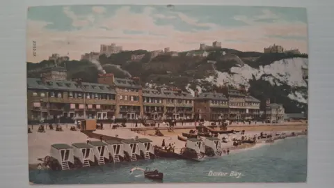 English Heritage A postcard with an illustration of a beach with buildings, the white cliffs of Dover and Dover Castle. The illustration is labelled Dover Bay.