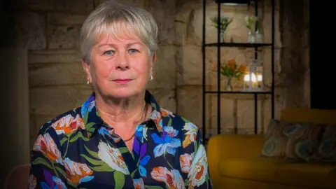 IWC Media Eileen Harrild, in a blouse of blue, red and white, is sitting in a room with stone walls and a yellow sofa in the background. She has short grey hair.