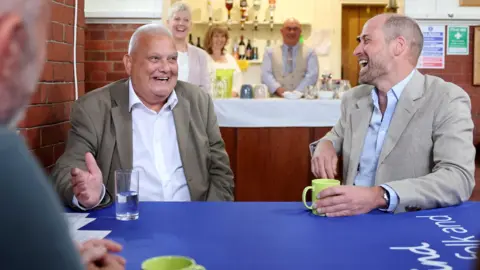 PA Media Prince William, to the right, and a man to the left sit at a table with a blue tablecloth. The man to the left has very short grey hair and is smiling, wearing a khaki suit jacket and white shirt. Prince William is turned to look at him and is laughing. He has his left hand around a green mug placed on the table and is wearing a beige jacket and pale blue shirt. 