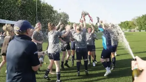 Andrew Harper Female football players celebrating on the pitch, spraying champagne and hugging.