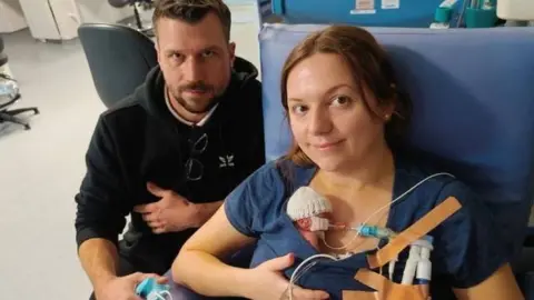 Family photo A woman with dark hair with a small baby on her chest. Behind her is a man with short hair and a beard, wearing a black hoody