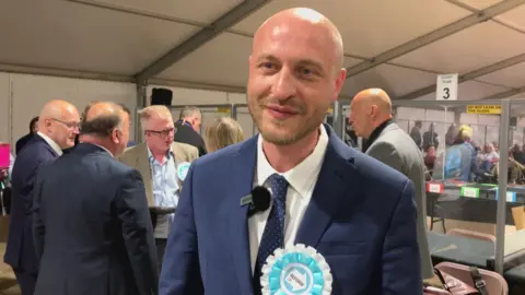 A smiling man with a bald head and wearing a blue suit and tie and Reform rosette.