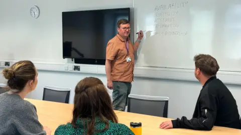 UEA Dr Mark Gregory, wears glasses, a brown shirt and jeans, and stands at a white board with a pen in a classroom. Two women and a man sit with their backs to the camera, looking at Dr Gregory.