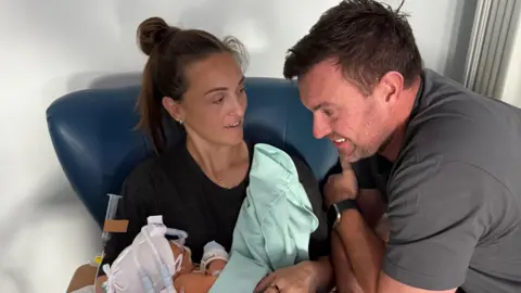 Jade Jade is seated in a hospital chair holding a baby wrapped in a light green blanket. The baby has medical tubes and monitoring devices attached. Chris is leaning close, resting an arm on the chair, with hands touching the seated person’s arm.