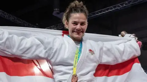 Getty Images Katie is also in a judo kit with a medal around her neck and holding an England flag out behind her. Her darker hair is tied up on her head.