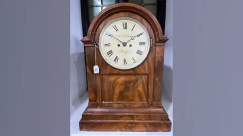 Chilcotts Auctioneers and Valuers A small brown clock with a white clock face and the numbers in roman numerals. The photo is on a light purple background.