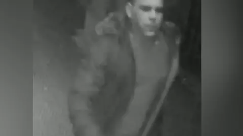 A blurred image of a man wanted in connection with a sexual assault in nightclub in Guildford, Surrey. He is wearing a dark puffer jacket.