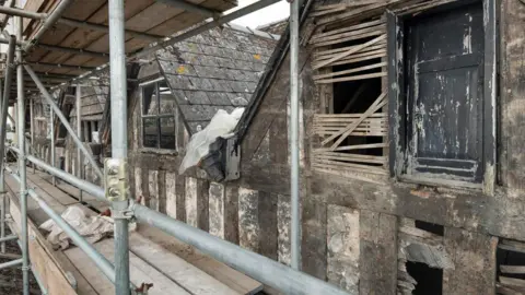 Historic England A similar angle to the first image, but in this case the building is in much worse shape. There is no yellow pigment or black paint. The building is surrounded by scaffolding and is covered in dilapidating wood.