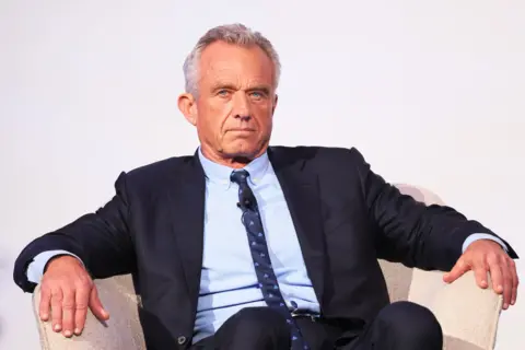 RFK Jr, wearing a suit, sits in an arm chair with his arms on the sides, with a neutral expression