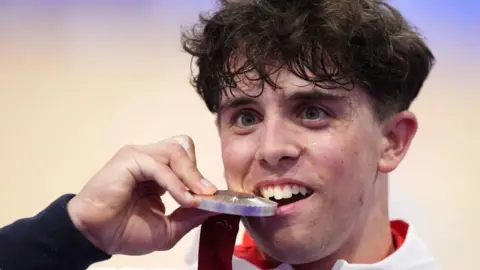 Great Britain's Archie Atkinson celebrates with the silver medal following the 4000m Individual Pursuit gold Final at the National Velodrome on day three of the Paris 2024 Summer Paralympics. He is biting the medal in celebration. 