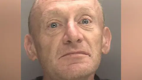 A mug shot of a man with very short light brown hair and facial stubble. He is wearing a dark coloured top