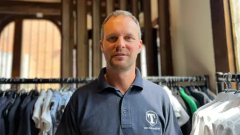 Becki Bowden/BBC A man with short grey hair and wearing a dark coloured polo shirt stands in a hall surrounded by clothes rails.