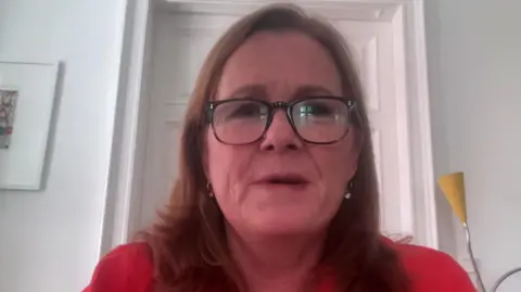 Kirsty Lang wearing a red top and classes is doing a zoom interview. There is a white door and wall behind her.