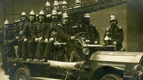 GMFRS A group of firefighters in matching uniforms wearing dated helmets and sat with ladders of an open-top old car. The photo is black and white and depicts the service in its earlier years.