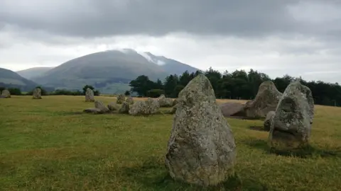 jackdog A part of the stone circle which is made up of large grey boulders of various shapes. There is a collection of smaller stones in the centre. The grass below is green with patches of yellow and brown. Behind it there is a group of trees with dark green leaves and a large fell to the centre-left. There are wispy clouds above the fell and the clouds further up are thicker and grey.