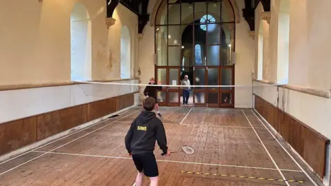 BBC Nave of church with wooden foor and tape setting out a court - a net is across the room with two boys in black shorts and tops holding badminton rackets playing a game.