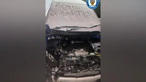 West Midlands Police A car that has been stripped of its bonnet and bumper. The windscreen has snow on it with the words "real grafter" written on it