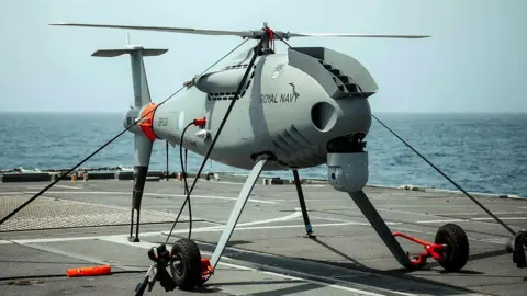Lt Cdr Fraser Simon A Peregrine drone is seen on the deck of a frigate, with a rotary blades on top and wheels beneath, it is grey with Royal Navy branding