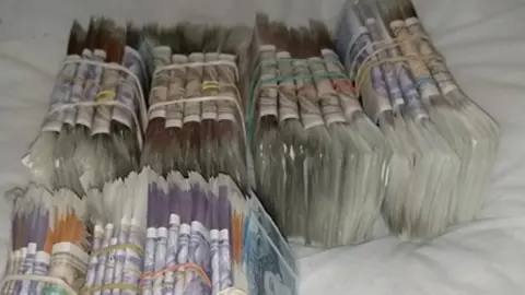 National Crime Agency Bank notes in the thousands are tied up and resting on what appears to be a bed.