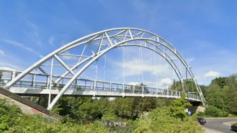 A white metal pedestrian bridge, which has a large arch over it, is suspended over a road in a residential area, surrounded by trees. There are cars coming off a roundabout to drive underneath it. The sky above is bright blue with a few wispy white clouds.