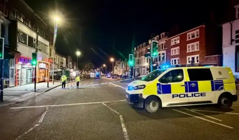 Leicester Media Online. Una furgoneta policial delante de la cinta policial en una carretera sin tráfico.