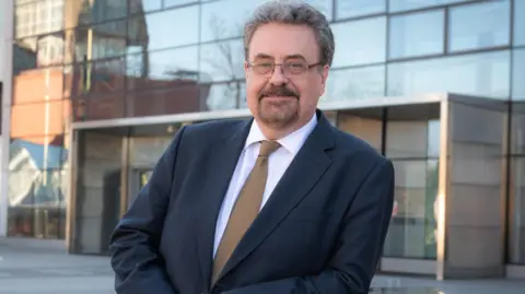 A man with grey hair,  a beard and glasses wearing a suit stands outside a large glass-fronted building.