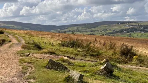 A country footpath alongside picturesque moorland and hills. Some rocks are in the foreground. 