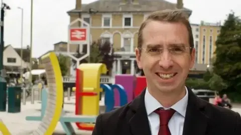Peter Taylor, wearing glasses, a dark suit and a red tie, stands in a public square near a railway station. Behind the person is a large yellow sculptural seating feature and colourful street furniture, with buildings, bicycles and a railway sign visible in the background.